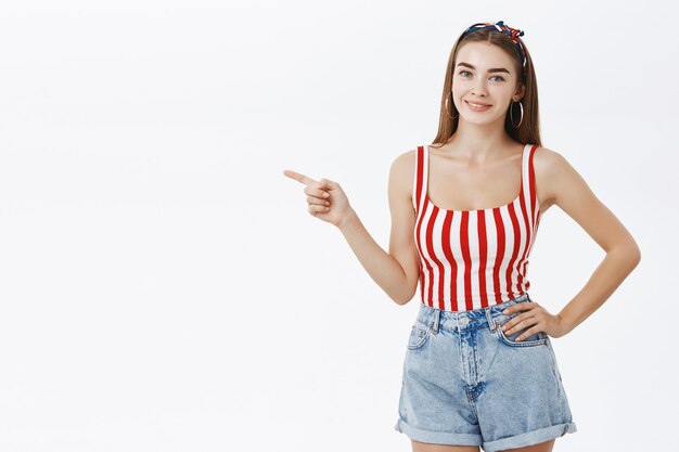 Portret van stijlvolle en vrouwelijke knappe vrouw in gestreepte pin-up top en korte broek met hand op taille glimlachend vriendelijk en wijzend naar links weergegeven: copyspace