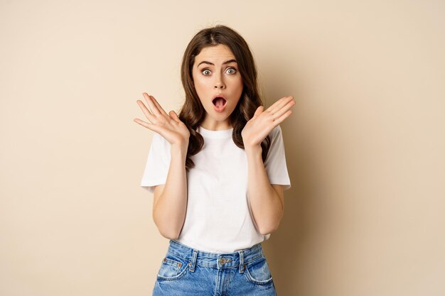 Portret van stijlvolle jonge vrouw hijgend, verbaasd en verbaasd kijkend, onder de indruk van iets, staande in t-shirt en spijkerbroek over beige achtergrond