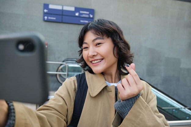 Portret van stijlvolle Koreaanse studente neemt selfie op straat, houdt smartphone vast en poseert op haar camera