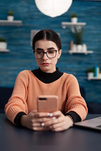 Portret van student met bril smartphone in handen houden. Influencer in thuiskantoor die e-mail leest op tochscreen smartphone. Freelancer werkt op afstand aan een bureau en sms't op sociale media.