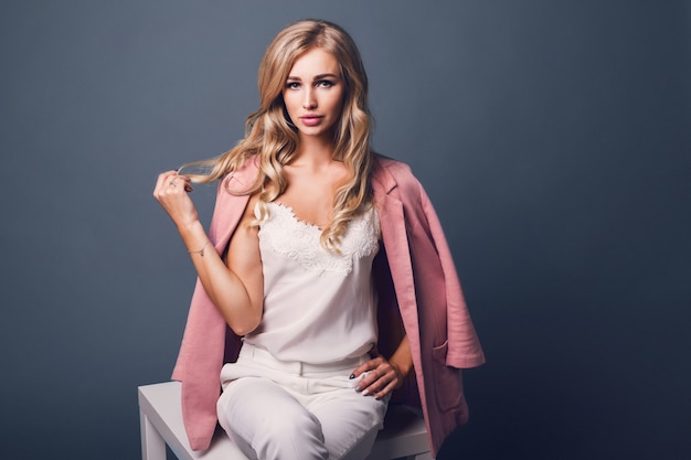 Portret van succesvolle jonge verleidelijke blonde vrouw in roze pastel jasje zittend op tafel