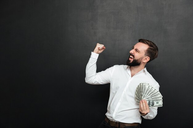 Portret van succesvolle man in wit shirt verheugend als winnaar met fan van 100 dollarbiljetten in de hand, balde vuist opzij over donkergrijs