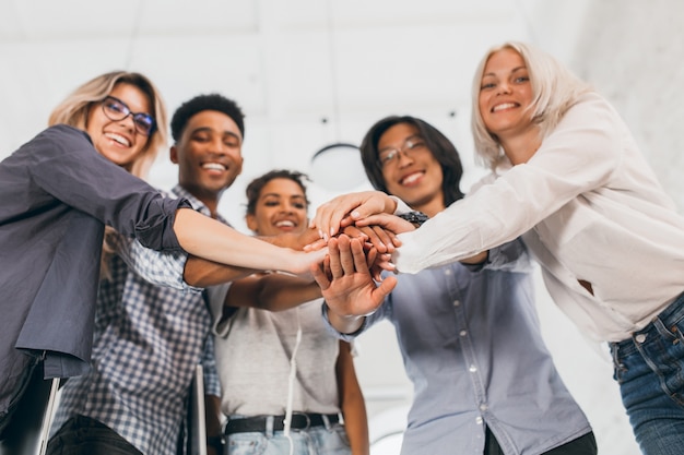 Portret van team van jonge kantoormedewerkers met hun handen in focus vervagen. Binnenfoto van lachende internationale studenten in stijlvolle kleding die elkaar ondersteunen voor examens.