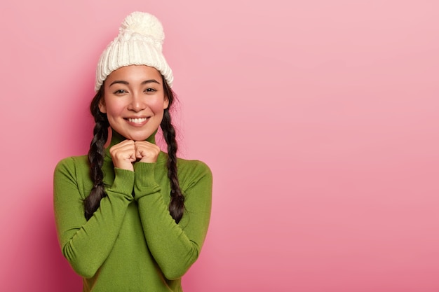 Portret van tedere mooie aziatische vrouw heeft rouge wangen, aangename glimlach, houdt de handen onder de kin, kijkt gelukkig direct naar de camera, draagt een gebreide muts