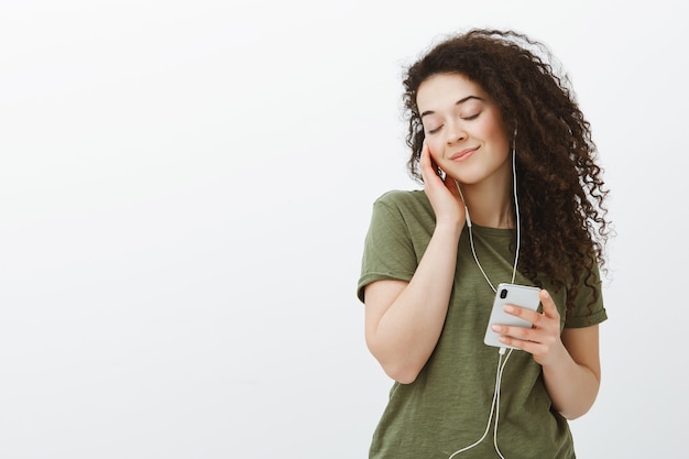 Portret van tedere mooie curly-haired brunette vrouw in casual outfit, ogen sluiten en gelukkig glimlachen, muziek in oortelefoons luisteren en smartphone houden