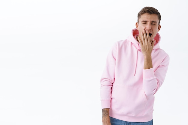 Portret van terughoudende knappe blonde man in roze hoodie, geeuwen met geopende mond en gesloten ogen