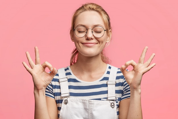 Gratis foto portret van tevreden blonde aantrekkelijke opgetogen vrouw geeft ok teken aan, houdt de ogen dicht, is in een goed humeur, blij om weekends te hebben, geïsoleerd over roze