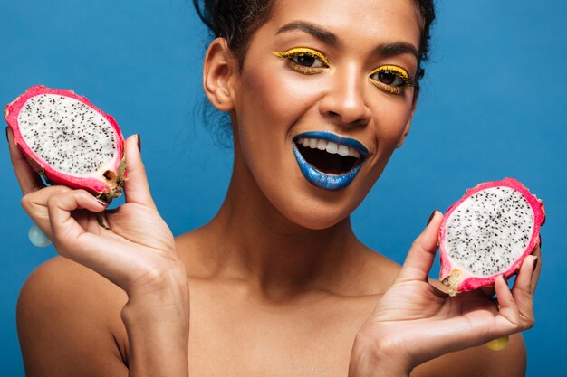 Portret van tevreden mulatvrouw die met heldere make-up van rijp pitayafruit genieten die in de helft over blauwe muur wordt gesneden