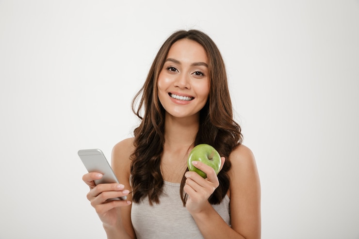 Gratis Foto portret van tevreden vrouw met perfecte glimlach gebruikend zilveren smartphone en etend verse groene appel geïsoleerd over witte muur