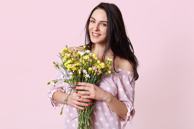 Portret van tevreden vrouwelijk model met donker haar, gekleed in polka dot jurk, glimlacht positief