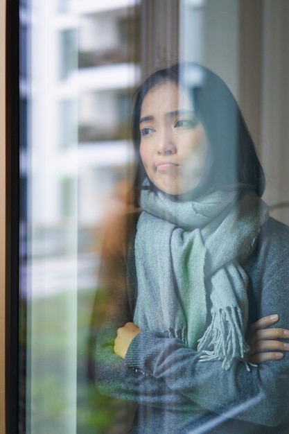 Gratis foto portret van trieste zieke koreaanse vrouw die uit het raam kijkt en thuis blijft omdat het coronavirus ziek is en het huis niet kan verlaten