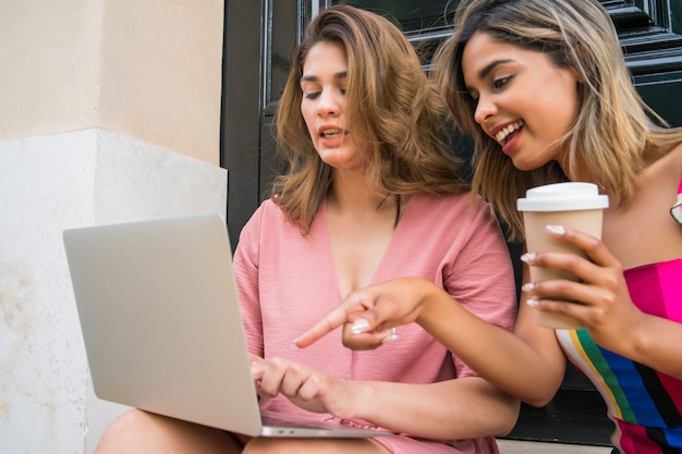 Portret van twee jonge vrienden die een laptop gebruiken en koffie drinken terwijl ze buiten zitten. Stedelijk begrip. Technologieconcept.