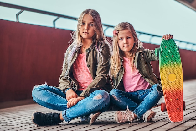 Portret van twee kleine zusjes gekleed in trendy kleding die samen op een skateboard bij een brugvoetgang zitten.