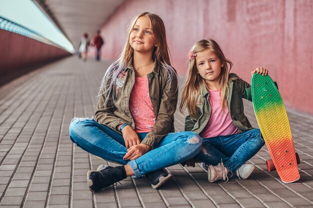 Portret van twee kleine zusjes gekleed in trendy kleding die samen op een skateboard bij een brugvoetgang zitten.