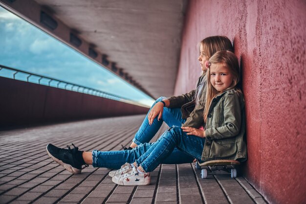 Portret van twee kleine zusjes gekleed in trendy kleding, leunend op een muur terwijl ze op een skateboard bij de brugvoet zitten.
