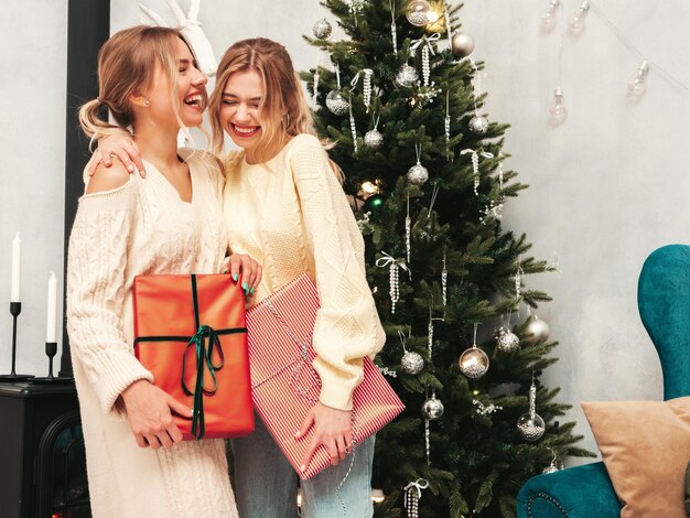 Portret van twee mooie blonde vrouwen Modellen poseren in de buurt van versierde kerstboom op oudejaarsavond Vrouw plezier klaar voor feest Vrienden gekleed in warme wintertruien