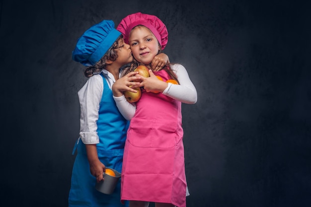 Portret van twee schattige kleine koks. Kleine jongen met bruin krullend haar gekleed in een blauw koksuniform en mooi schoolmeisje gekleed in een roze koksuniform, knuffelen en kussen samen terwijl hij kookt