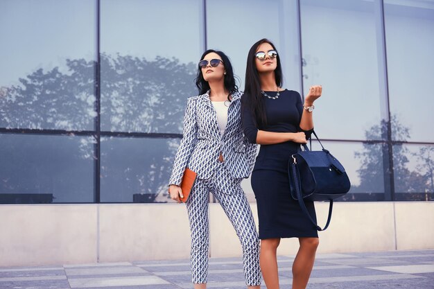 Portret van twee zakenvrouwen gekleed in stijlvolle formele kleding, staande in een centrum tegen een achtergrond van wolkenkrabber.