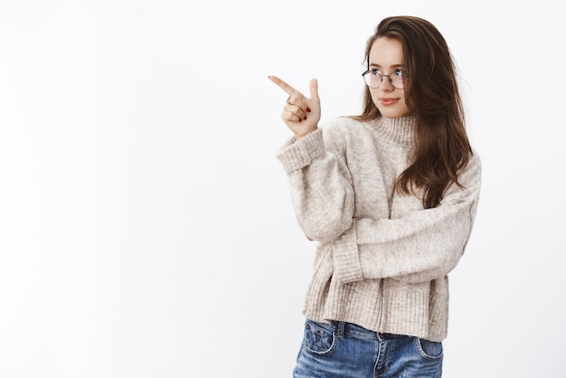 Portret van vastberaden slimme en aantrekkelijke charmante brunette vrouw in bril en trui die aangeeft naar links te kijken met nieuwsgierigheid en verlangen over grijze muur