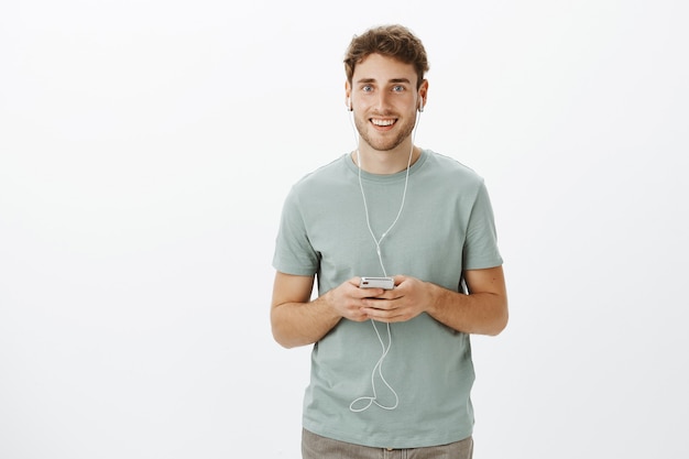 Portret van verbaasd gelukkig knappe man met baard en blond haar, smartphone vasthouden en oortelefoons dragen terwijl u naar muziek luistert, genietend van geweldig geluid en kwaliteit van oordopjes