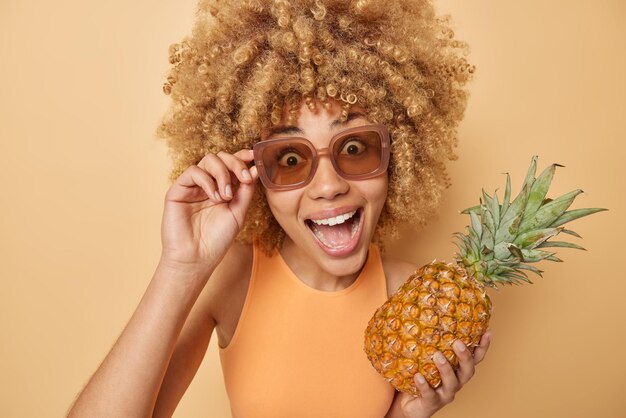 Portret van verbaasde gelukkige jonge vrouw met krullend borstelig haar draagt een zonnebril houdt verse ananas vol vitamines kijkt graag naar camera houdt mond geopend poses tegen beige achtergrond