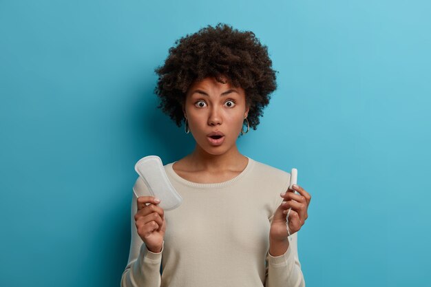 Portret van verbijsterd emotionele jonge Afro-Amerikaanse vrouw houdt verschillende soorten producten voor vrouwelijke hygiëne maandverband en tampon, draagt casual witte trui, heeft menstruatie of menstruatie