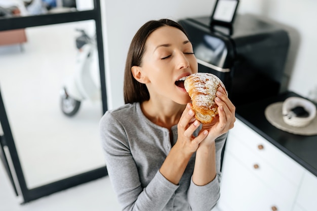 Portret van verheugende vrouw eet lekkere croissant thuis