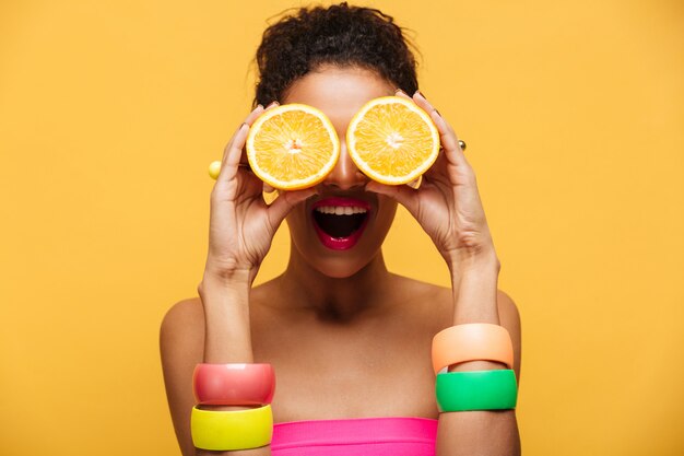 Portret van vermakelijke Afro-Amerikaanse vrouw met mode-accessoires plezier en die de ogen bedekt met twee geïsoleerde helften van sinaasappel, over gele muur