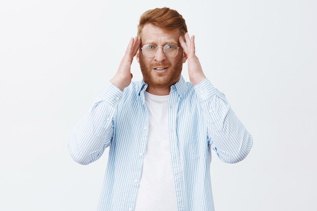 Portret van vermoeide knappe mannelijke ondernemer met gember haar en baard in glazen, hand in hand op tempels en ongericht staren, hoofdpijn hebben