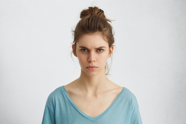 Portret van verontwaardigde jonge vrouw met ovaal gezicht, blauwe ogen en haarbroodje die blauwe toevallige sweater dragen die haar wenkbrauwen fronsen die met iets ontevreden zijn.