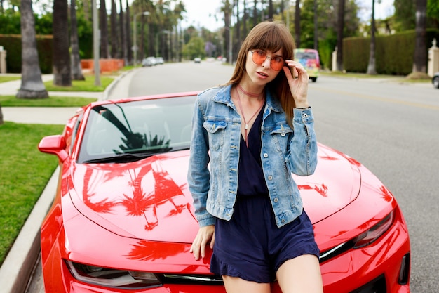 Portret van verrast brunette vrouw zittend op de motorkap van geweldige rode cabriolet sport auto in Californië close-up.
