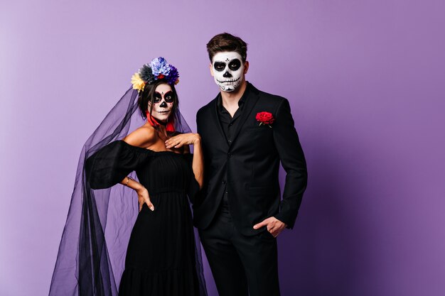 Portret van verraste dame in beeld van bruid voor Halloween en haar vriendje in klassiek kostuum met geschilderd gezicht in vorm van schedel.