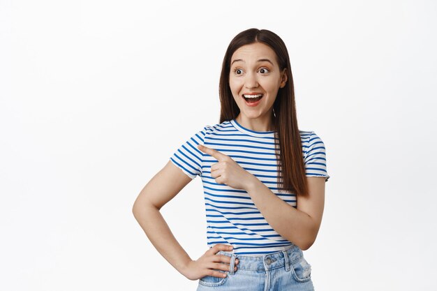 Portret van verraste, gelukkige jonge brunette vrouw die uitverkoop checkt, met de vinger wijst en links naar banner kijkt, geamuseerd glimlacht, staande tegen een witte achtergrond