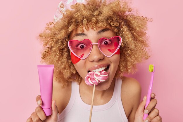 Portret van verraste vrolijke vrouw met krullend borstelig haar houdt lolly in mond houdt tandpasta en tandenborstel past schoonheidspleisters toe onder ogen draagt roze hartzonnebril poseert binnen