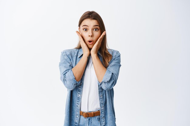 Portret van verraste vrouw hoort groot nieuws, hijgend en ontroerend gezicht geschokt, verbaasd starend, staande in vrijetijdskleding over wit