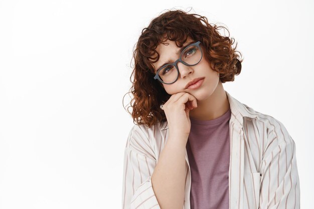 Portret van verveelde krullende studente met een bril, leunend op de hand en kijkend met onverschilligheid, staren verveeld en terughoudend, luisterend naar iets saai en oninteressant, witte achtergrond.