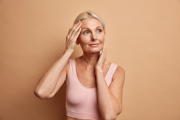 Portret van volwassen oudere Europese vrouw raakt gezicht zachtjes heeft perfecte huid en kijkt bedachtzaam weg geniet van haar zachte teint geeft om uiterlijk tevreden na anti-verouderingsprocedure