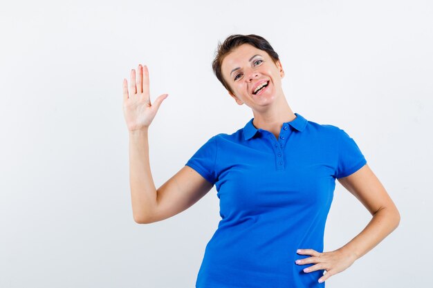 Portret van volwassen vrouw zwaaiende hand om afscheid te nemen in blauw t-shirt en op zoek blij vooraanzicht