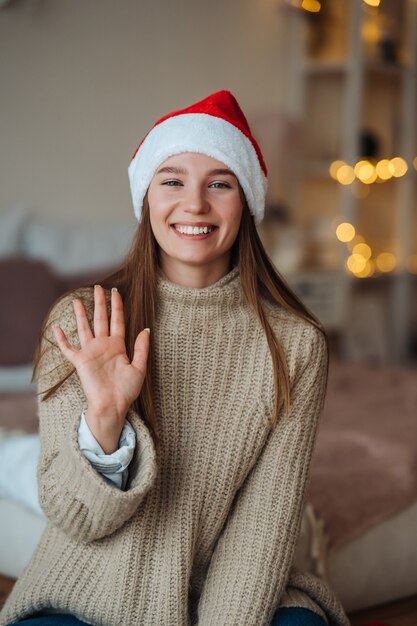 Portret van vriendelijke vriendelijke brunette vrouw in kerstmuts opgeheven hand zwaaien en hallo zeggen tegen camera, genieten van kersttijd.