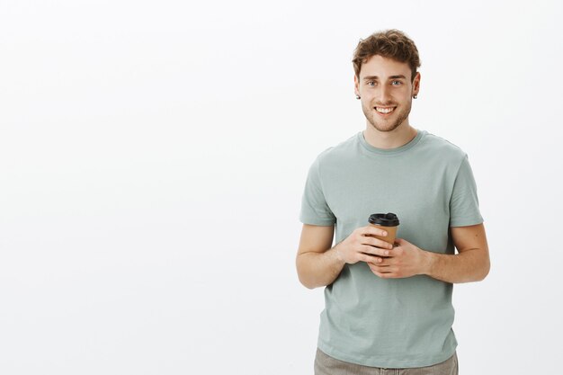 Portret van vriendelijke zelfverzekerde jonge ondernemer met blond haar en oorbellen, kopje koffie te houden en vreugdevol glimlachen terwijl terloops praten met collega