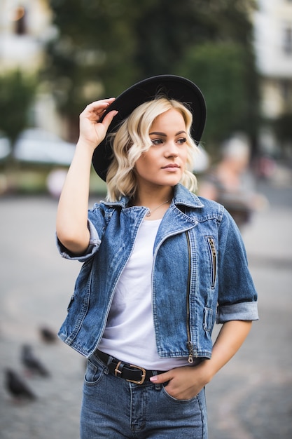 Portret van vrij blond meisje in stadscentrum gekleed in jeansreeks en wit t-shirt en zwarte hoed