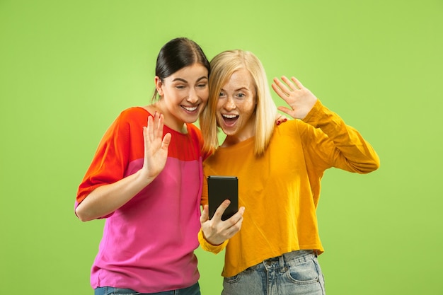 Portret van vrij charmante meisjes in casual outfits geïsoleerd op groene studio achtergrond. Vriendinnen of lesbiennes praten over smartphone. Concept van LGBT, gelijkheid, menselijke emoties, liefde, relatie.