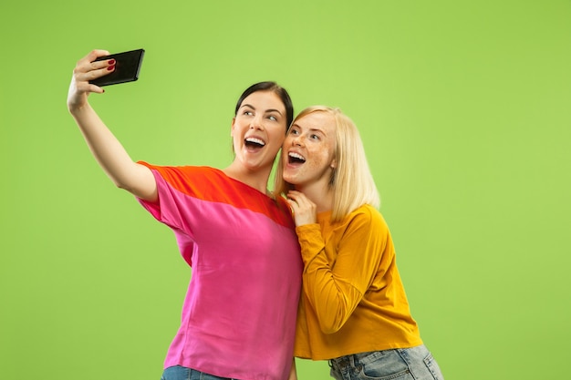 Portret van vrij charmante meisjes in casual outfits geïsoleerd op groene studio achtergrond. Vriendinnen of lesbiennes selfie maken. Concept van LGBT, gelijkheid, menselijke emoties, liefde, relatie.