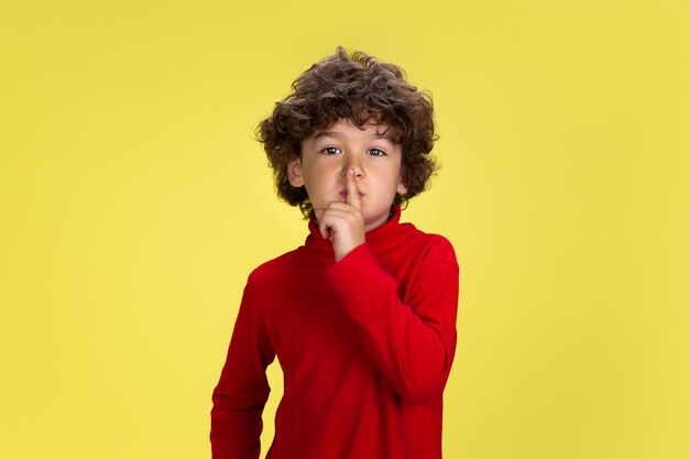 Portret van vrij jonge krullende jongen in rode slijtage op gele studioachtergrond. Jeugd, expressie, onderwijs, leuk concept. Peuter met heldere gezichtsuitdrukking en oprechte emoties.