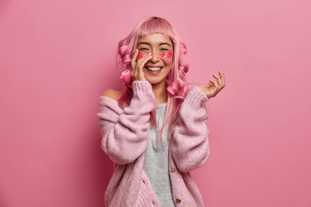 Portret van vrolijk Aziatisch meisje gebruikt hydrogel patches met anti-rimpeleffect, draagt haarkrulspelden op roze haar, glimlacht oprecht, draagt casual trui