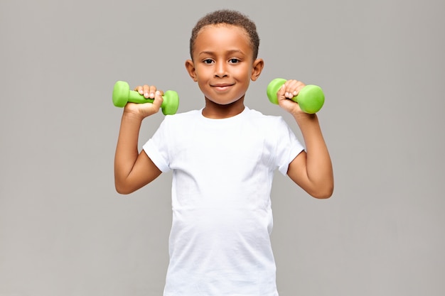 Portret van vrolijke Afro-Amerikaanse jongen met magere armen glimlachend gelukkig tijdens het trainen in de sportschool met twee halters, sterk, gezond atletisch lichaam gaan bouwen. Fitness en kinderen