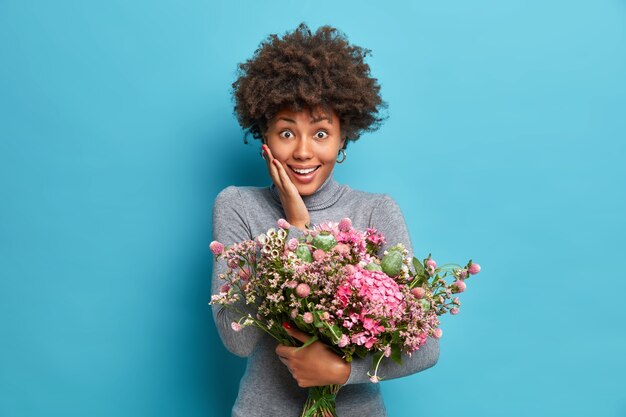 Portret van vrolijke Afro-Amerikaanse vrouw accepteert gefeliciteerd met verjaardag ontvangt bloemen heeft uitdrukking verbaasd