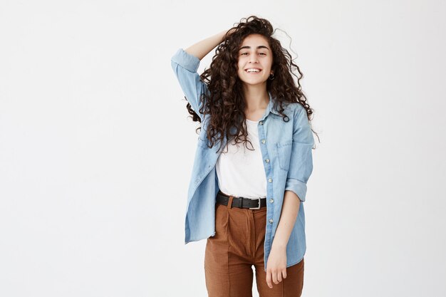 Portret van vrolijke brunette met lang golvend haar, gekleed in denim overhemd en bruine broek, blij goed nieuws te ontvangen, Student meisje lacht breed, verheugt het leven, drukt positieve emoties uit