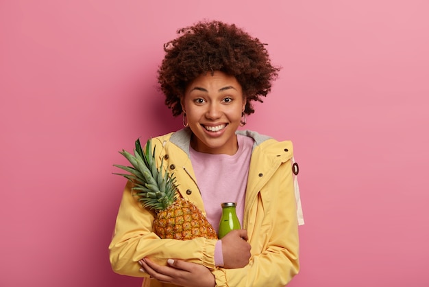 Portret van vrolijke gekrulde vrouw omarmt verse, sappige ananas en groene smoothie gemaakt van fruit, draagt gele anorak, glimlacht positief