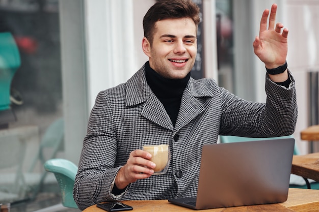 Portret van vrolijke jonge kerel glimlachende en golvende hand die benoeming met vriend in straatkoffie hebben